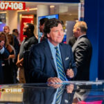 Christian Petition EXPLODES Over Tucker Carlson Guest… News anchor sitting at desk with people applauding.