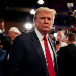 Trump–MAMDANI Whiplash Turns Ugly… A serious-looking man in a suit with a red tie surrounded by a crowd and cameras