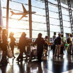 DHS Could HALT Flights to Sanctuary Cities… People queuing inside airport terminal with airplane flying outside.