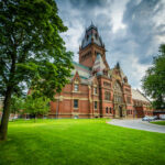 Red-brick Gothic-style building with tall clock tower beside a green lawn