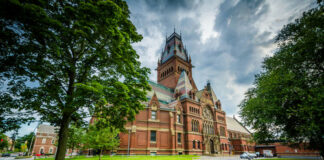 Red-brick Gothic-style building with tall clock tower beside a green lawn
