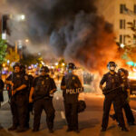 Police officers in riot gear facing a chaotic protest scene with smoke and fire