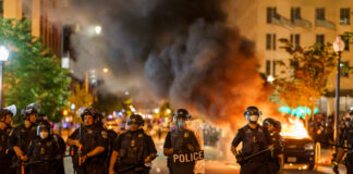 Police officers in riot gear facing a chaotic protest scene with smoke and fire