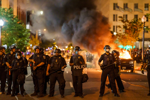 Police officers in riot gear facing a chaotic protest scene with smoke and fire