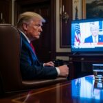 A political figure seated in the Oval Office, watching a television broadcast