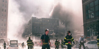 Firefighters walking through debris near a heavily damaged smoking building