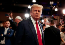 Peace Tease: Trump Works the War Phones A serious-looking man in a suit with a red tie surrounded by a crowd and cameras