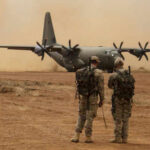 Two soldiers watch a military transport plane on a dusty airstrip