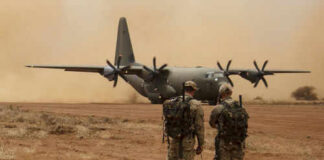 Two soldiers watch a military transport plane on a dusty airstrip