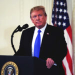 A man in a suit standing at a podium with the presidential seal, in front of an American flag