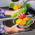 Hands washing cucumbers and bell peppers under a kitchen faucet