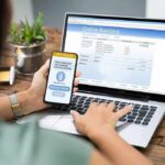 Person using smartphone to approve bank transfer beside laptop with online banking