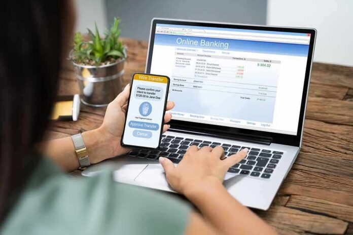 Person using smartphone to approve bank transfer beside laptop with online banking