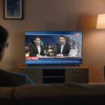 Person watching a live news broadcast on TV in a cozy living room