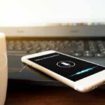 Smartphone with voice assistant on screen beside laptop and coffee mug
