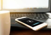 Smartphone with voice assistant on screen beside laptop and coffee mug