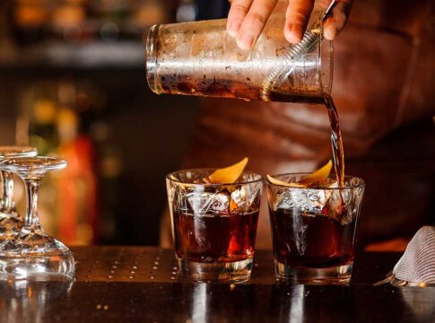 Pouring cocktails into two glasses with lemon twists.