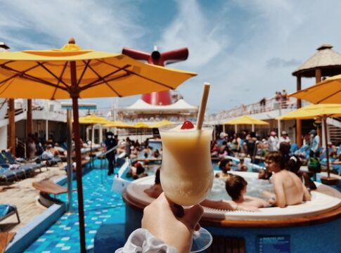 A hand holding a tropical drink by a pool on a cruise ship