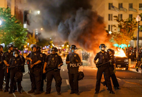 Police officers in riot gear facing a chaotic protest scene with smoke and fire